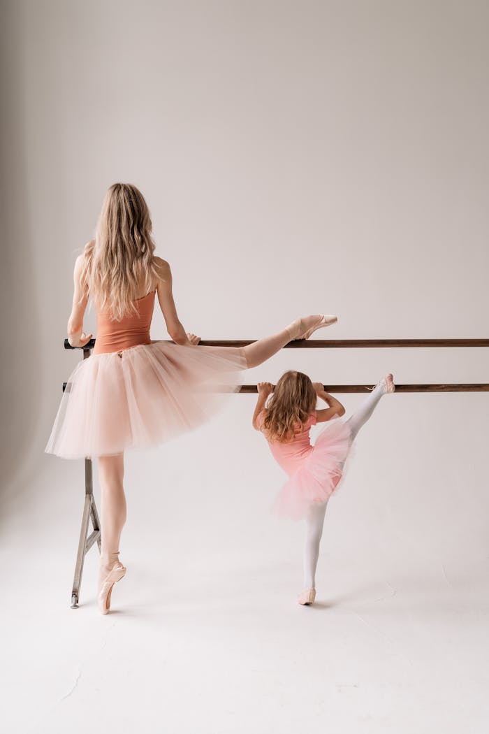 A mother and daughter in tulle skirts practicing ballet at a barre in a studio.