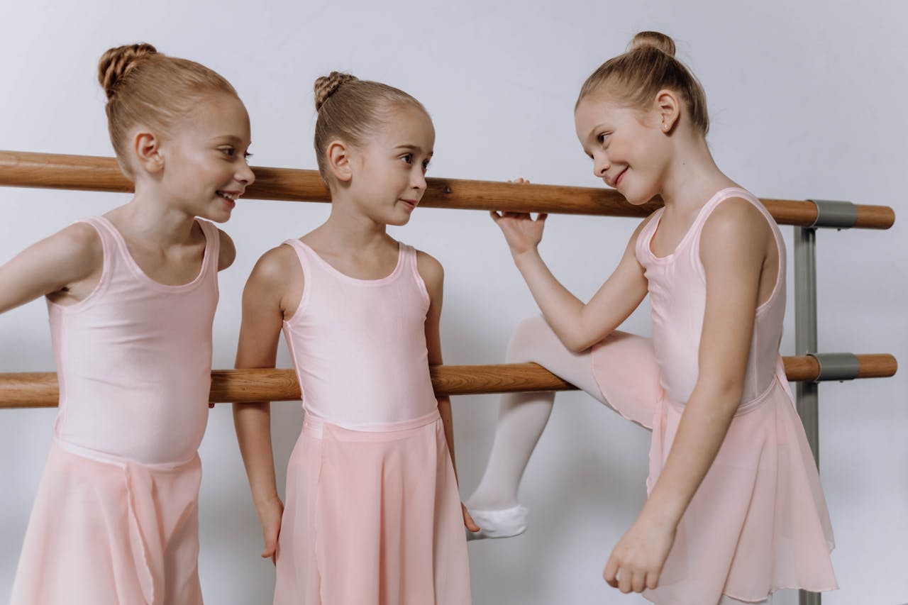 Three young girls in ballet attire share smiles during a ballet class.
